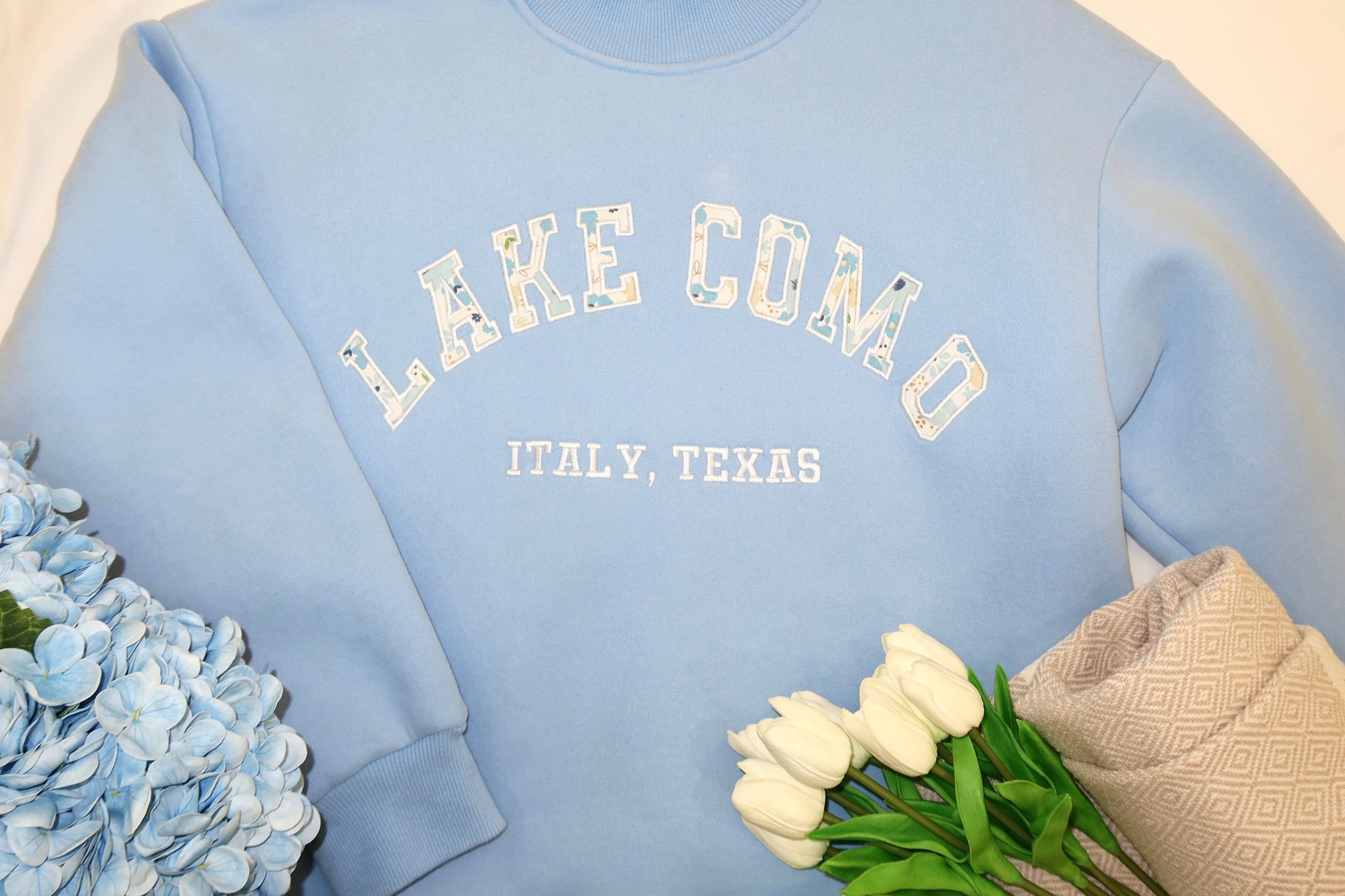 Light blue sweatshirt with 'Lake Como' text, placed on a beige surface with flowers.